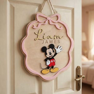 Op de afbeelding: Houten deurbord met een roze strik en rand, met de naam "Liam James" in gouden letters en een Mickey Mouse-afbeelding. Het bord is ontworpen om aan een deur te hangen.
