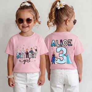 May include: Pink short-sleeved t-shirt with a graphic featuring characters and text from the movie "Frozen". The front says "Birthday girl" and the back says "ALICE 3". The shirt is worn by a young child.
