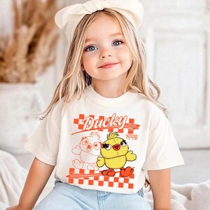 Może przedstawiać: Kremowa koszulka z grafiką z kreskówki przedstawiającą kaczkę, napisem "Ducky" i rokiem "1995" w kolorze pomarańczowym. Projekt zawiera wzór w szachownicę. Koszulka jest noszona przez małe dziecko.