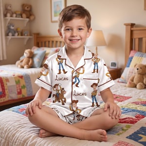 May include: A white short-sleeved pajama set featuring a repeating pattern of the character Woody from Toy Story and the name Lucas printed in brown script. The button-down shirt has a notched collar, brown piping, and a single chest pocket.