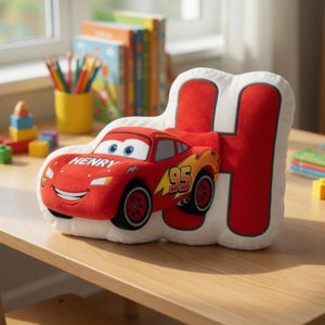 May include: A plush pillow in the shape of the letter "H" with a red race car design. The car is red with the number "95" and the name "Henry" printed on it. The pillow is on a wooden table.