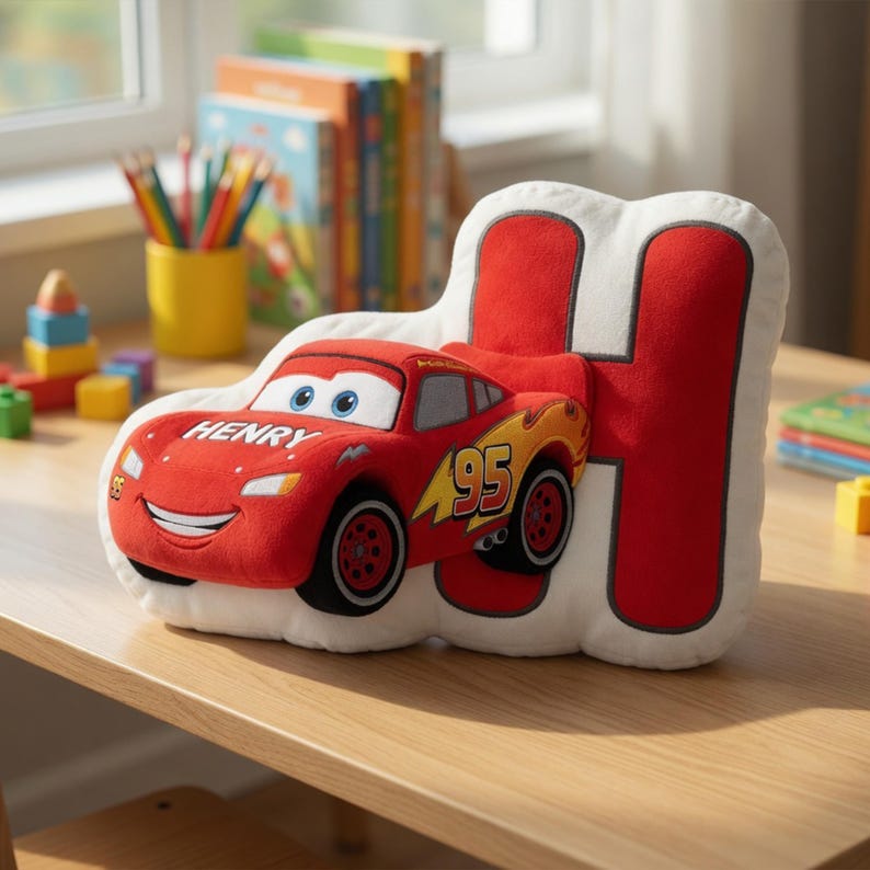 May include: A plush pillow in the shape of the letter "H" with a red race car design. The car is red with the number "95" and the name "Henry" printed on it. The pillow is on a wooden table.