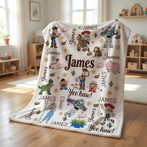 Puede incluir: Una manta de felpa blanca con personajes de dibujos animados y el nombre "James" en marr&oacute;n. La manta tiene ilustraciones de personajes, estrellas y cactus. Tambi&eacute;n se incluye la frase "Yee haw!"