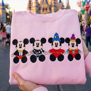May include: A pale pink sweatshirt with four Mickey Mouse designs in various outfits. The cartoon characters are in black, red, and yellow. The background is blurred, showing a castle and people.