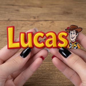Può includere: Una toppa in tessuto ricamato che riporta il nome Lucas in lettere gialle con un bordo rosso spesso. A destra del nome si trova un piccolo e dettagliato personaggio ricamato che raffigura un cowboy con cappello marrone, camicia gialla e fazzoletto rosso.