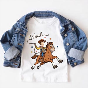 以下が含まれることがあります： 茶色の馬に乗ったカウボーイの漫画が描かれた白いTシャツ。カウボーイの上には「Noah」という名前が筆記体で書かれ、デザインの周りにはカラフルな星が散りばめられています。青いデニムジャケットがTシャツの上にかけられています。