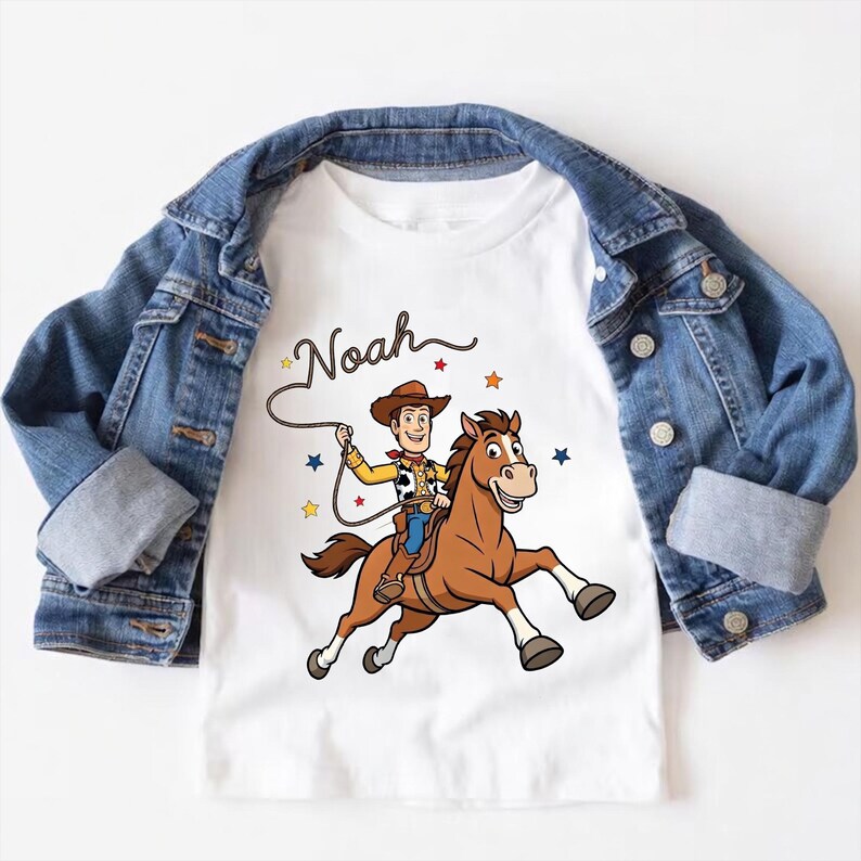 以下が含まれることがあります： 茶色の馬に乗ったカウボーイの漫画が描かれた白いTシャツ。カウボーイの上には「Noah」という名前が筆記体で書かれ、デザインの周りにはカラフルな星が散りばめられています。青いデニムジャケットがTシャツの上にかけられています。