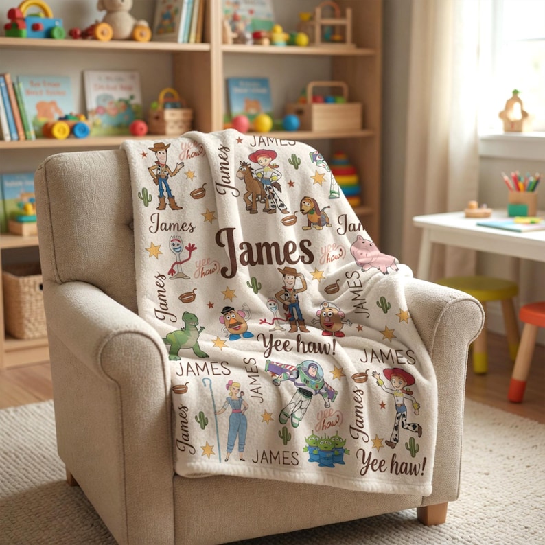 Puede incluir: Una manta crema con personajes de Toy Story, el nombre "James" y las frases "Yee haw!" y "Howdy". La manta est&aacute; sobre un sill&oacute;n beige en una habitaci&oacute;n infantil, con una estanter&iacute;a al fondo.