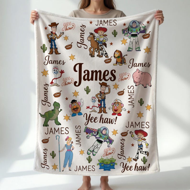 Puede incluir: Una manta de felpa blanca con personajes de dibujos animados y el nombre "James" en una fuente grande y marr&oacute;n. La manta tiene varios personajes de dibujos animados, estrellas y cactus. La frase "Yee haw!" tambi&eacute;n es visible.