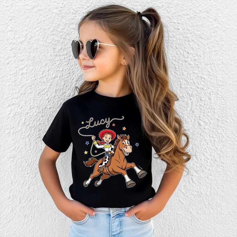 以下が含まれることがあります： 茶色の馬に乗ったカウガールの漫画が描かれた黒いTシャツ。グラフィックの上には「Lucy」という名前があります。カウガールは赤い帽子をかぶり、ロープを持っています。星が画像を囲んでいます。Tシャツは若い女の子が着ています。