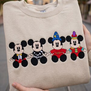 May include: Beige crew neck sweatshirt with four Mickey Mouse character designs. Each character is dressed in a different outfit, including a magician's hat and a circus-style outfit. The sweatshirt is held by a person.