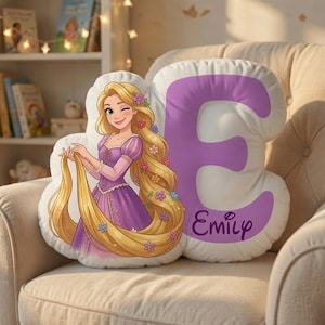 May include: A white, cloud-shaped throw pillow featuring a cartoon illustration of a blonde character with long hair wearing a purple dress, alongside a large purple letter E and the name Emily printed in a cursive purple font on the pillow.