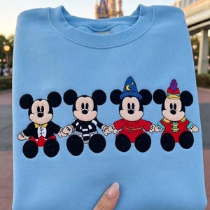 May include: Pale blue sweatshirt with four Mickey Mouse designs in various outfits. The characters are holding hands and are in black, red, and white. A castle and monorail are visible in the background.
