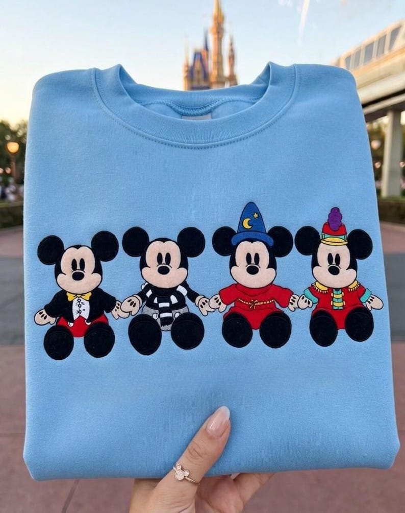 May include: Pale blue sweatshirt with four Mickey Mouse designs in various outfits. The characters are holding hands and are in black, red, and white. A castle and monorail are visible in the background.