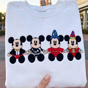 May include: White sweatshirt with four Mickey Mouse character designs. Each Mickey is dressed in a different outfit, including a tuxedo, striped shirt, wizard attire, and a festive ensemble. The designs are in black, red, yellow, and blue.