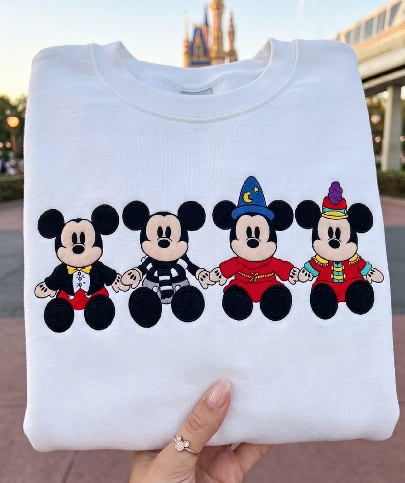 May include: White sweatshirt with four Mickey Mouse character designs. Each Mickey is dressed in a different outfit, including a tuxedo, striped shirt, wizard attire, and a festive ensemble. The designs are in black, red, yellow, and blue.