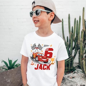 May include: White t-shirt featuring a red cartoon race car, checkered flags, the number 6, and the name "JACK" in red. The shirt is a birthday theme, suitable for a child.
