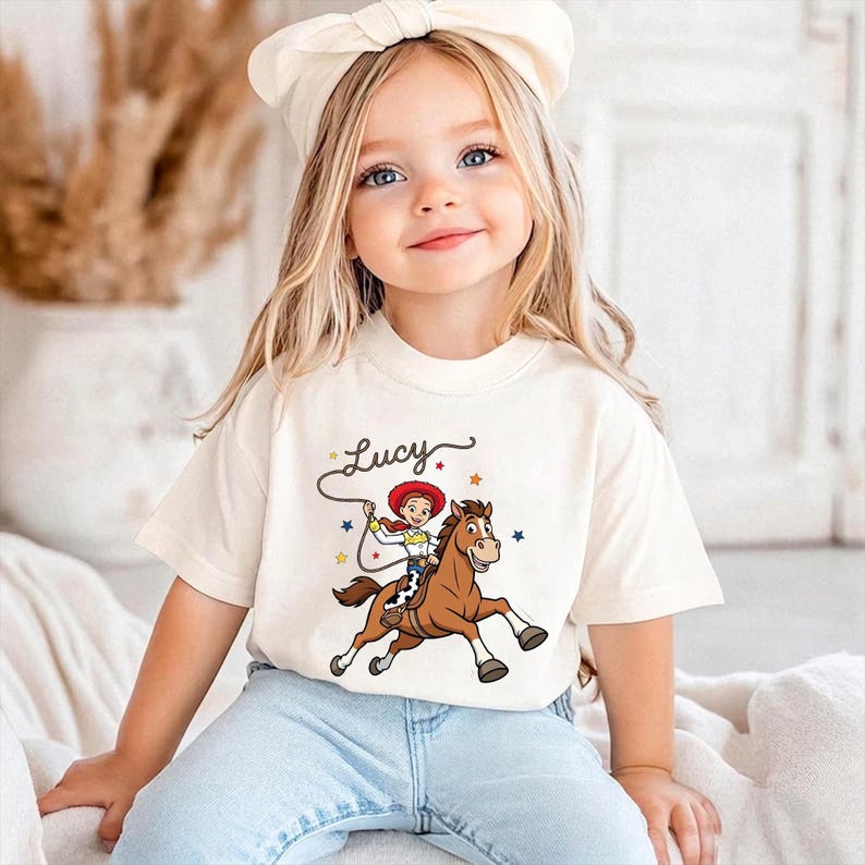 以下が含まれることがあります： クリーム色のTシャツ。茶色の馬に乗ったカウガールの漫画イラストが描かれており、グラフィックの上には「Lucy」という名前があります。カウガールは赤い帽子をかぶり、ロープを持っています。このシャツは青いジーンズと組み合わせています。