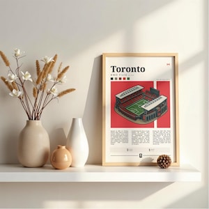Peut inclure: Une affiche encadrée représentant le stade BMO Field de Toronto. L'affiche a un fond rouge avec le stade au centre. Le mot "Toronto" est en haut, avec le drapeau canadien. L'affiche est exposée sur une étagère blanche, avec des vases.