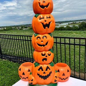 Op de afbeelding: Een stapel van vijf oranje plastic pompoenen met uitgesneden gezichten, op elkaar gestapeld. Twee kleinere pompoenen en een skelet hand liggen voor de stapel. De pompoenen staan op een groene basis.