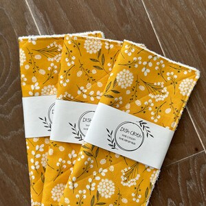 May include: Three yellow dishcloths with a white floral pattern and white stitching around the edges. The dishcloths are stacked on top of each other. The top dishcloth has a white label that says "Dish Cloth 100% cotton 100% handmade".
