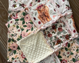 May include: A patchwork baby quilt with a floral pattern in shades of pink, white, and burgundy. The quilt features a square panel with a brown and white Highland cow and calf illustration. The quilt is made with soft, quilted fabric and has a ruffled edge.