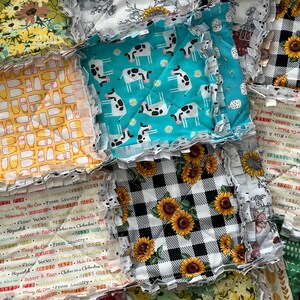 May include: A patchwork quilt made of various fabric squares. The squares feature different patterns, including cows, sunflowers, checkered patterns, and a farmhouse scene. The quilt is made with a frayed edge, giving it a rustic and handmade look.