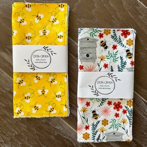 May include: Two handmade dishcloths, one with a yellow background and a bee pattern, the other with a white background and a floral pattern with bees. Both cloths have the text "Dish Cloth 100% Cotton 100% Handmade".