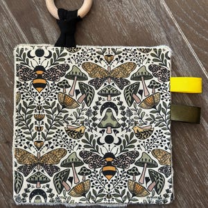 May include: A white fabric baby sensory toy with a black and white bee, butterfly, and mushroom pattern. The toy has a wooden ring attached to a black fabric loop and three fabric tags in yellow, green, and black.