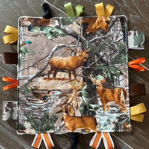 May include: A square baby security blanket with a camouflage pattern featuring deer and ducks. The blanket has various colored ribbons and loops around the edges, including orange, brown, green, and yellow. The overall design is a woodland theme.