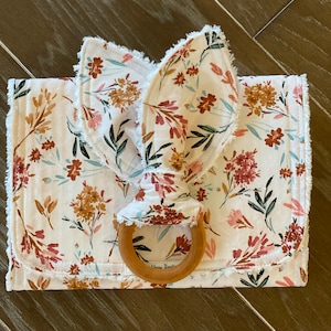 May include: A white muslin baby blanket with a floral print and a wooden teething ring attached. The blanket has a frayed edge and the ring has the words "Moon Bean" inscribed on it.