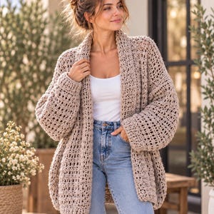 May include: A beige open-front cardigan featuring a loose, textured crochet knit pattern with long, wide sleeves and a shawl collar, worn over a plain white tank top and light blue denim jeans with a visible metal button and zipper fly closure.