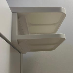 May include: A white, wall-mounted shelf unit with two rectangular shelves. The shelves have rounded corners and are mounted at a right angle to the wall. The unit is made of a smooth, solid material and is designed for storage or display.