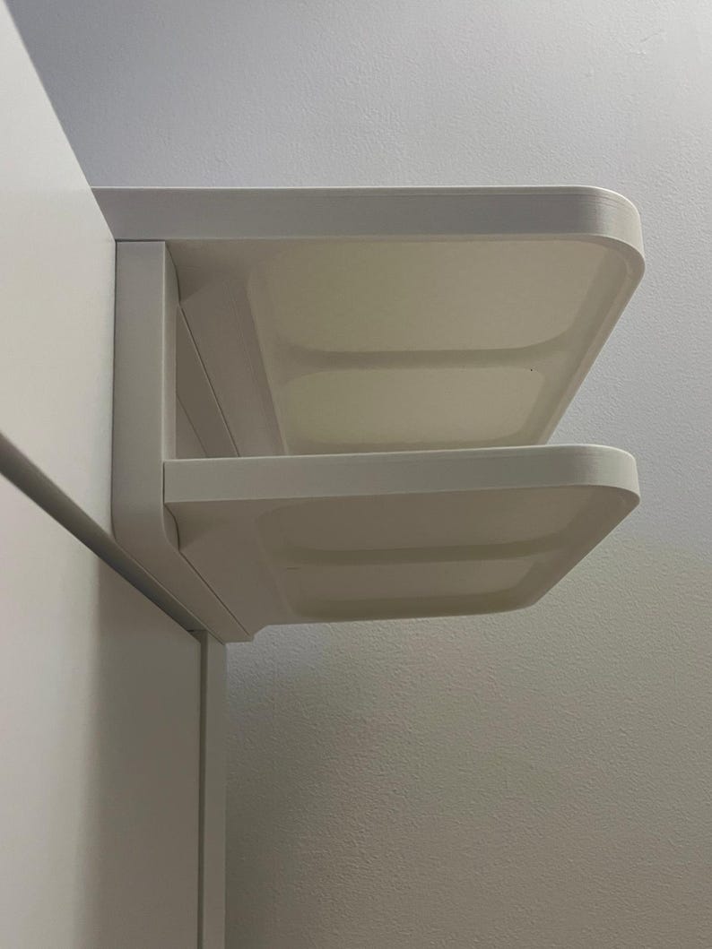 May include: A white, wall-mounted shelf unit with two rectangular shelves. The shelves have rounded corners and are mounted at a right angle to the wall. The unit is made of a smooth, solid material and is designed for storage or display.