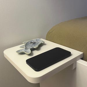 May include: A white bedside table with a rounded edge, holding a black smartphone and a ceramic fish-shaped dish. The dish is light blue and contains a silver ring. The table is attached to a white bed frame.