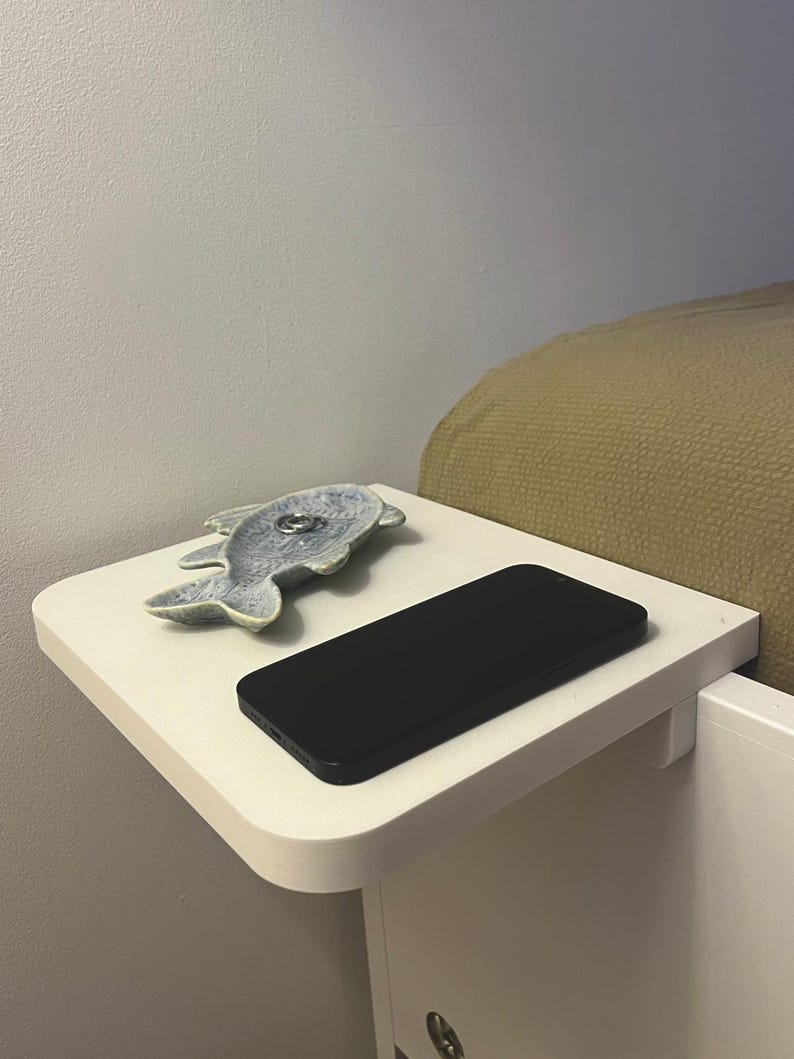 May include: A white bedside table with a rounded edge, holding a black smartphone and a ceramic fish-shaped dish. The dish is light blue and contains a silver ring. The table is attached to a white bed frame.