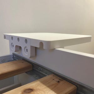 May include: A white, rectangular bed shelf with rounded corners is mounted on a bed frame. The shelf is supported by a white bracket with multiple holes and screws. Below the shelf, two wooden bed slats are visible.