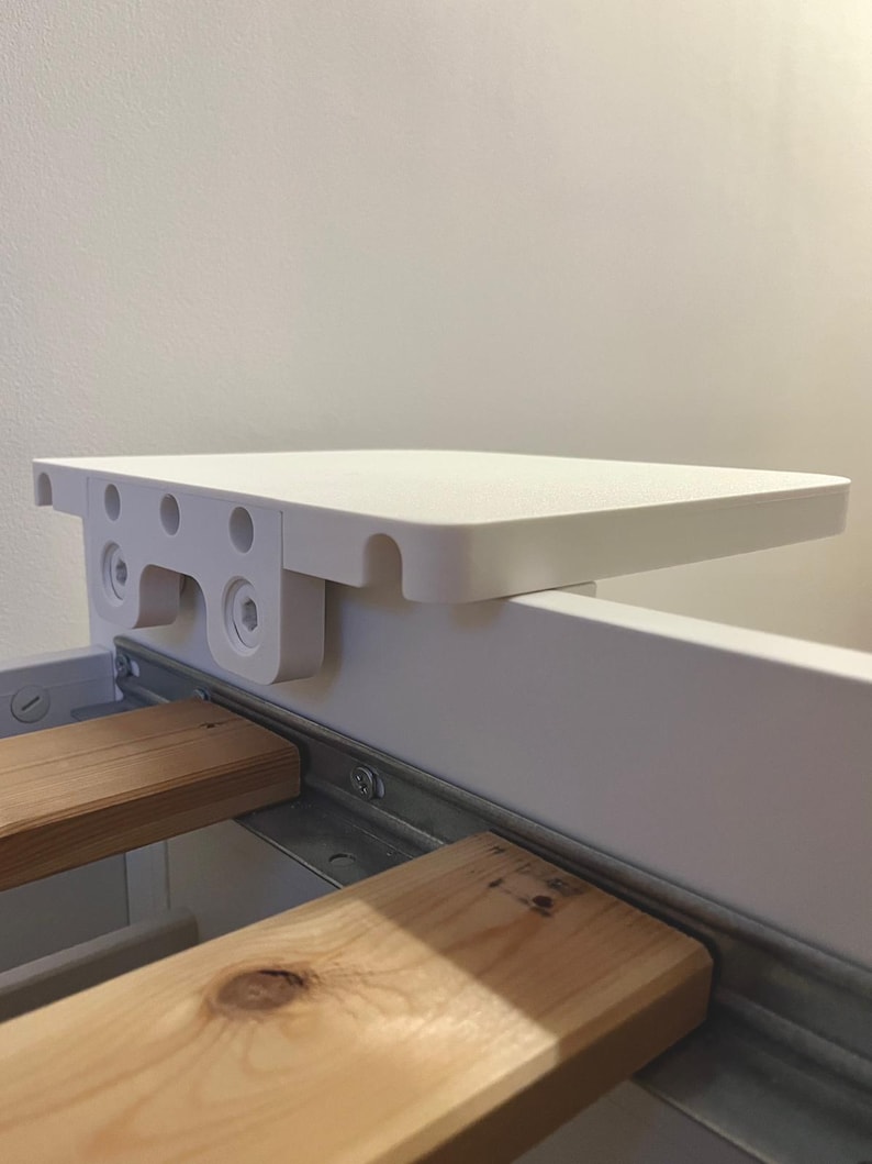 May include: A white, rectangular bed shelf with rounded corners is mounted on a bed frame. The shelf is supported by a white bracket with multiple holes and screws. Below the shelf, two wooden bed slats are visible.
