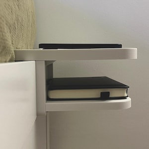 May include: A white bedside shelf attached to a bed frame. A black smartphone rests on the top shelf, and a black notebook with a cream-colored edge sits on the lower shelf. The shelf is a simple, modern design.