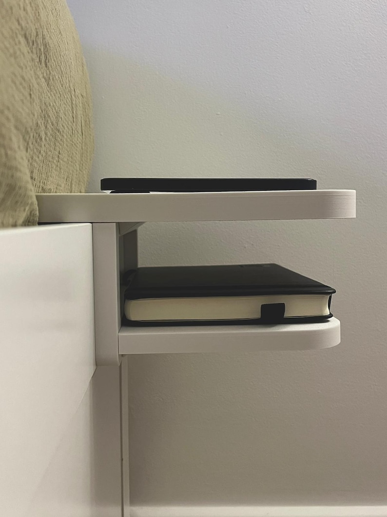 May include: A white bedside shelf attached to a bed frame. A black smartphone rests on the top shelf, and a black notebook with a cream-colored edge sits on the lower shelf. The shelf is a simple, modern design.