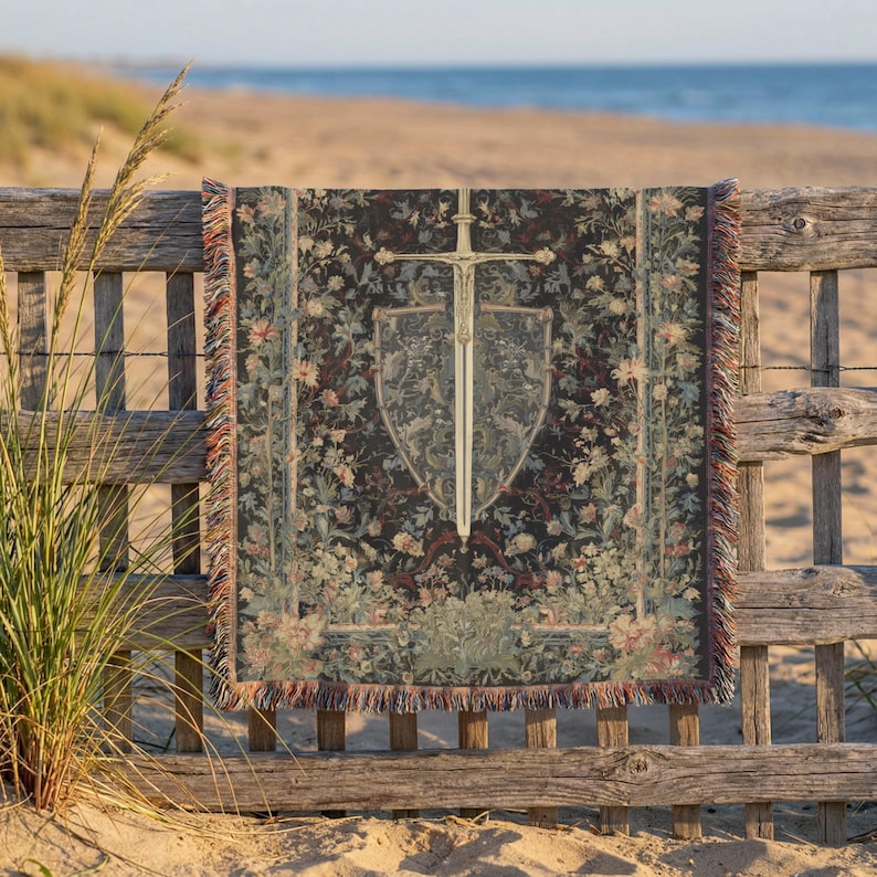 May include: A tapestry throw blanket with a medieval design featuring a sword and shield. The blanket has a dark background with floral patterns in shades of green, red, and beige. The fringe is red and beige. The blanket is displayed on a wooden fence.