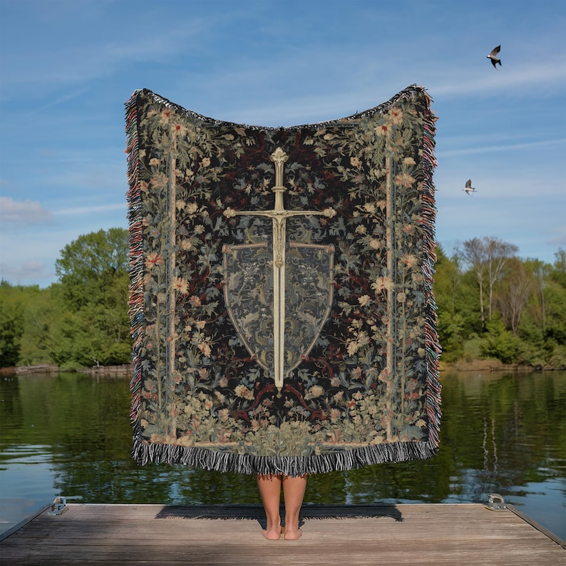 May include: A tapestry blanket featuring a sword and shield design. The blanket has a dark background with floral patterns and a fringed edge. The sword and shield are centered on the blanket, with a detailed design. The blanket is held up by a person standing on a wooden dock.