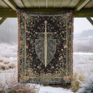 May include: A tapestry blanket with a medieval sword and shield design. The blanket features a dark background with floral patterns in shades of green, red, and beige. The sword and shield are in a light gold color, creating a striking contrast.