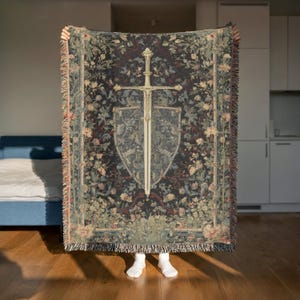 May include: A tapestry-style throw blanket with a dark background and floral patterns. The center features a sword and shield design in cream and gray. The blanket has fringed edges.