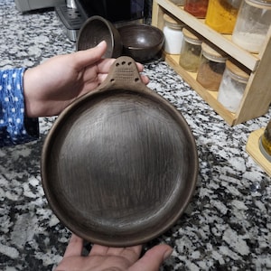 Puede incluir: Un plato de cerámica marrón oscuro hecho a mano con una superficie texturizada y un asa con un diseño rallado. El plato es redondo y parece ser parte de un juego de utensilios de cocina. Otros cuencos de cerámica son visibles en el fondo.