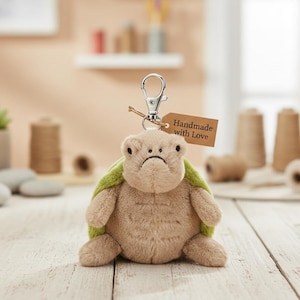 May include: A plush turtle keychain with a sad expression and a green shell. The keychain has a silver clasp and a small tag that reads "Handmade with Love". The turtle is made of soft, tan-colored fabric.