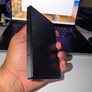 May include: A black, rectangular object with a triangular top is held in a hand. The object appears to be a case or container. The background includes a laptop displaying a screen with the letters "STL".