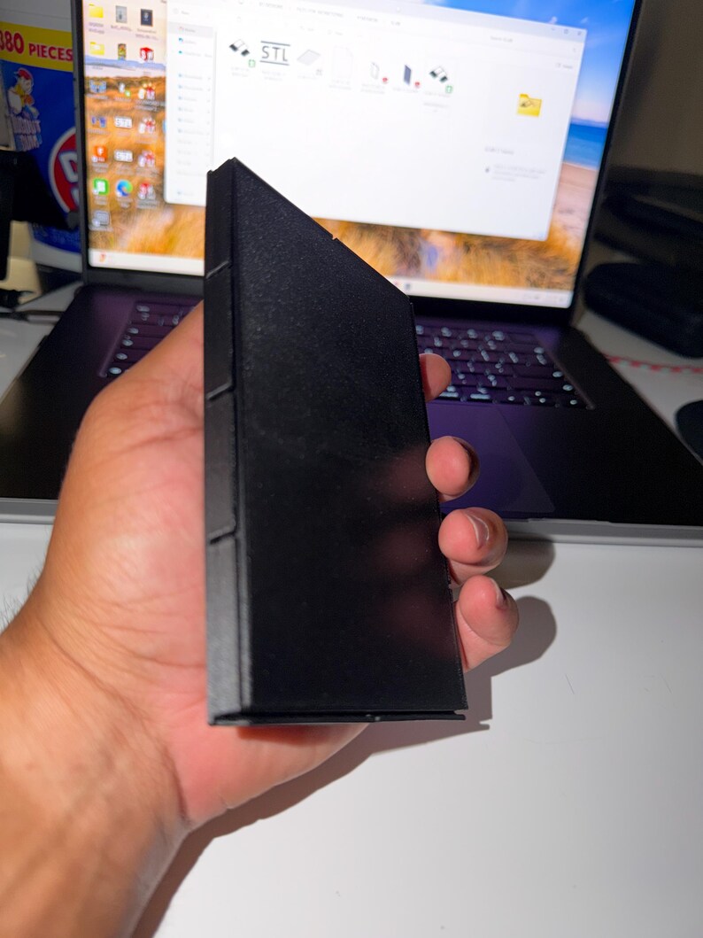 May include: A black, rectangular object with a triangular top is held in a hand. The object appears to be a case or container. The background includes a laptop displaying a screen with the letters "STL".