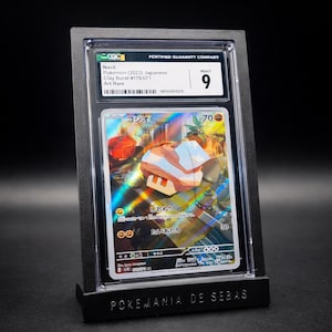 May include: A graded Pok&eacute;mon card featuring a holographic illustration of a character with Japanese text. The card is encased in a black holder with a "9" grade from the grading company. The holder is displayed on a black stand with the text "POKEMANIA DE SEBAS".