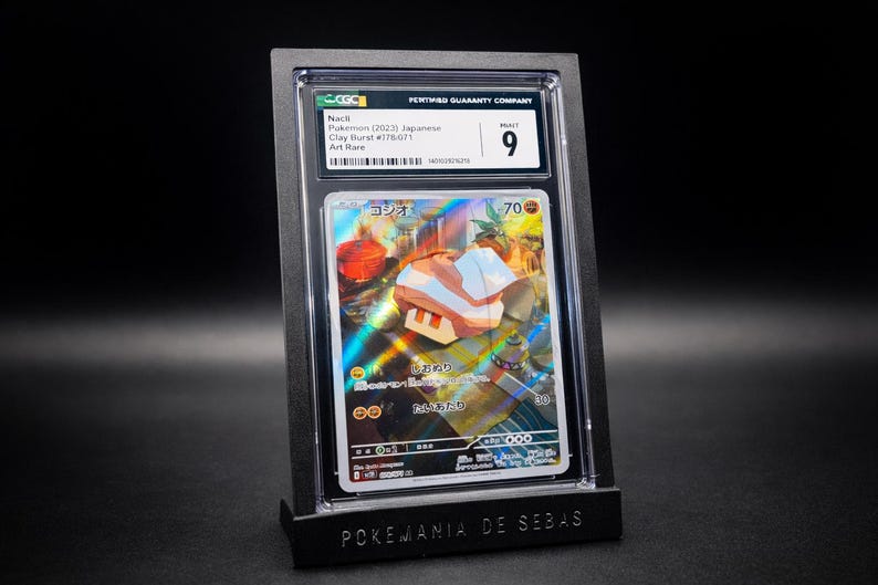 May include: A graded Pok&eacute;mon card featuring a holographic illustration of a character with Japanese text. The card is encased in a black holder with a "9" grade from the grading company. The holder is displayed on a black stand with the text "POKEMANIA DE SEBAS".